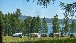 Un campamento con tiendas de campaña y vehículos junto a un lago, rodeado de árboles y colinas.