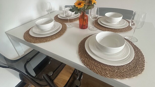 Ein Esstisch ist gedeckt mit weißen Tellern, Schüsseln und Gläsern, einer Blumenvase und gewebten Platzsets.