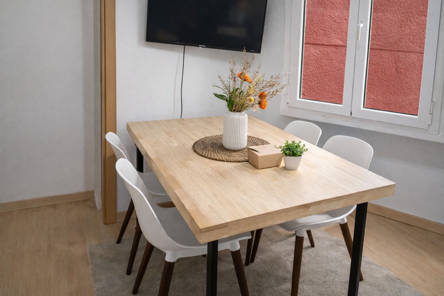En matplats med ett träbord, vita stolar, en vas med blommor, en krukväxt och en tv på väggen.