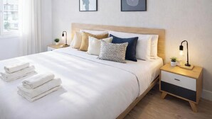 Uma cama bem arrumada com lençóis brancos e toalhas dobradas, uma cabeceira de madeira, mesas de cabeceira com abajures e vista para o exterior por uma janela.