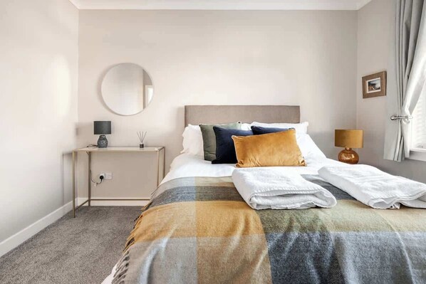 A neatly arranged hotel room with a bed, bedside table, lamp, and a round mirror on the wall. - Coorie Doon, Largs, spacious 2 bedroom apartment (North Ayrshire Council)