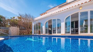 Ein Swimmingpool mit klarem, blauem Wasser, umgeben von einem weißen Gebäude mit großen Glasüren und einem Ziegeldach.