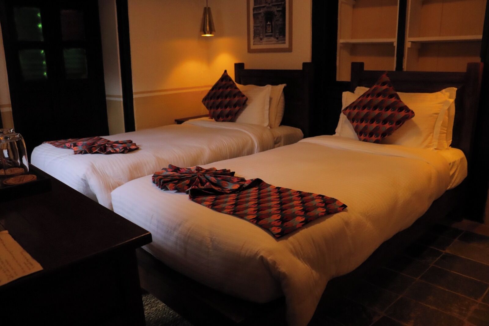 A hotel room with two beds, a bedside table with a lamp, and a framed picture on the wall.