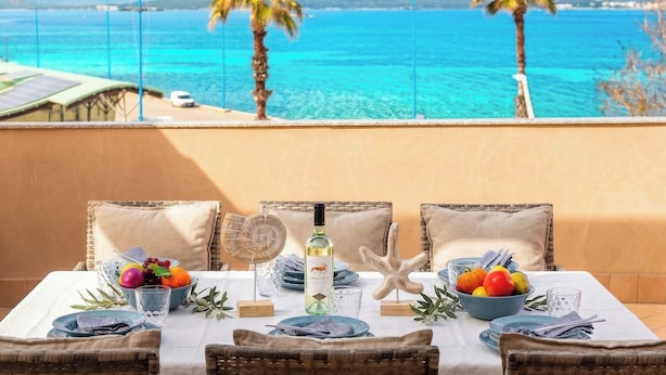 A table set with plates, glasses, and a bottle, overlooking a coastal view with palm trees and mountains. - Sunset Harbor Penthouse with a sun-drenched panoramic terrace and garage (Alghero)