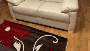 Een beige leren bank in een woonkamer met een rood en zwart tapijt, twee vloerlampen en een televisie op een rode metalen standaard.