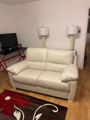 Un sofá de cuero color beige en una sala de estar con un tapete rojo y negro, dos lámparas de pie y un televisor sobre un soporte de metal rojo.