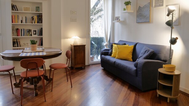A living room with a sofa, a dining table with chairs, a bookshelf, and a window with a view. - La Finestra sul Fiume - 5 min from the Basilica (Vicenza)