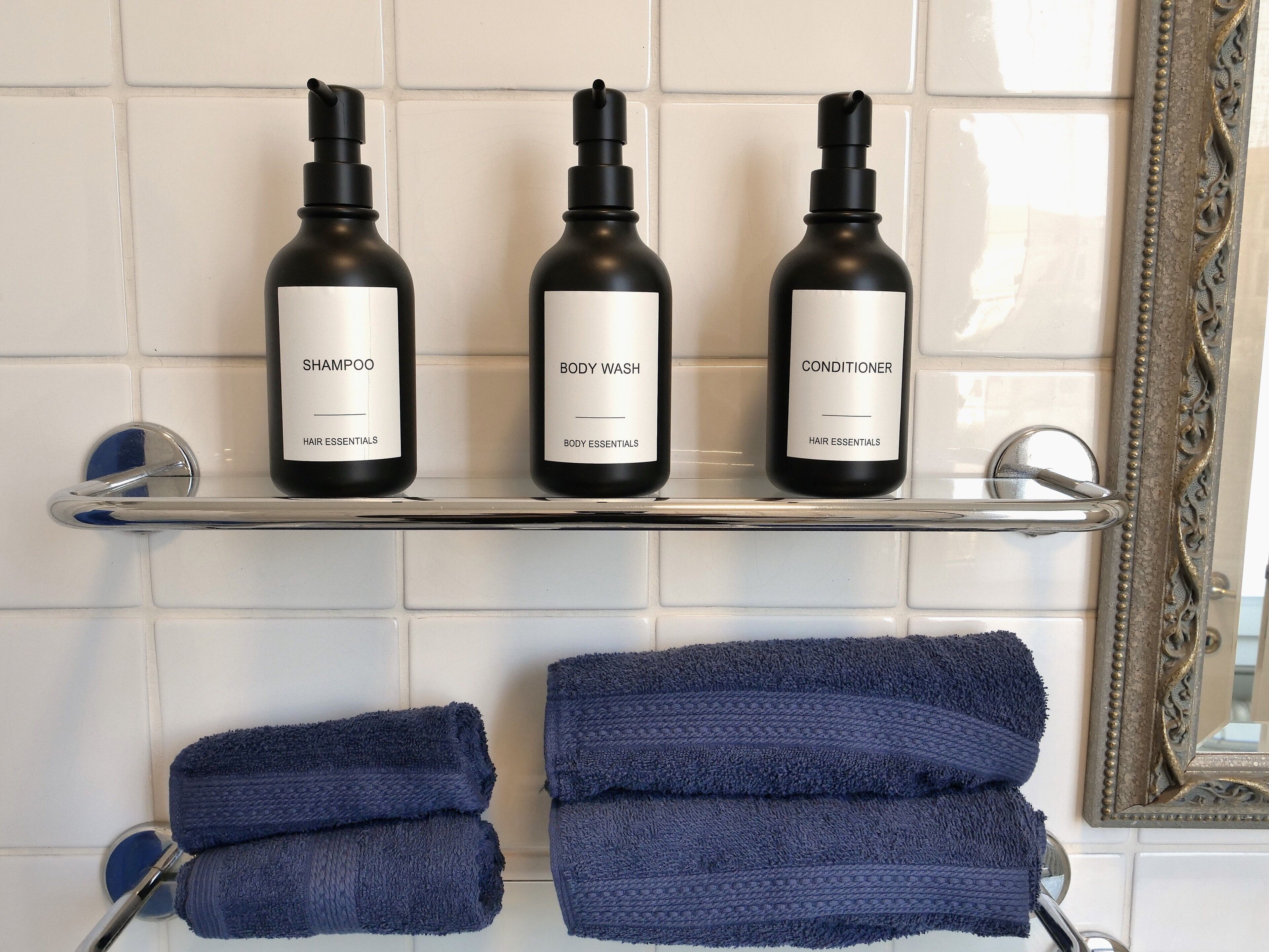 Kylpyhuoneen hyllyllä on kolme pulloa, joissa on etiketit: shampoo, suihkugeeli ja hoitoaine, sekä kaksi taitettua pyyhettä.
