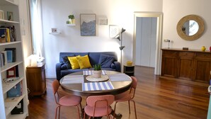 A living room with a round table, two chairs, a sofa with cushions, a bookshelf, a cabinet, and a mirror on the wall. - La Finestra sul Fiume - 5 min from the Basilica (Vicenza)
