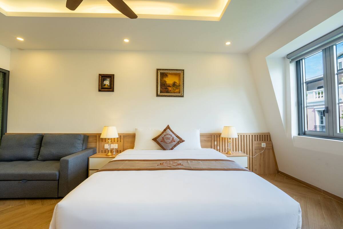 A hotel room with a bed, a sofa, two wall-mounted lamps, a ceiling fan, and a window.