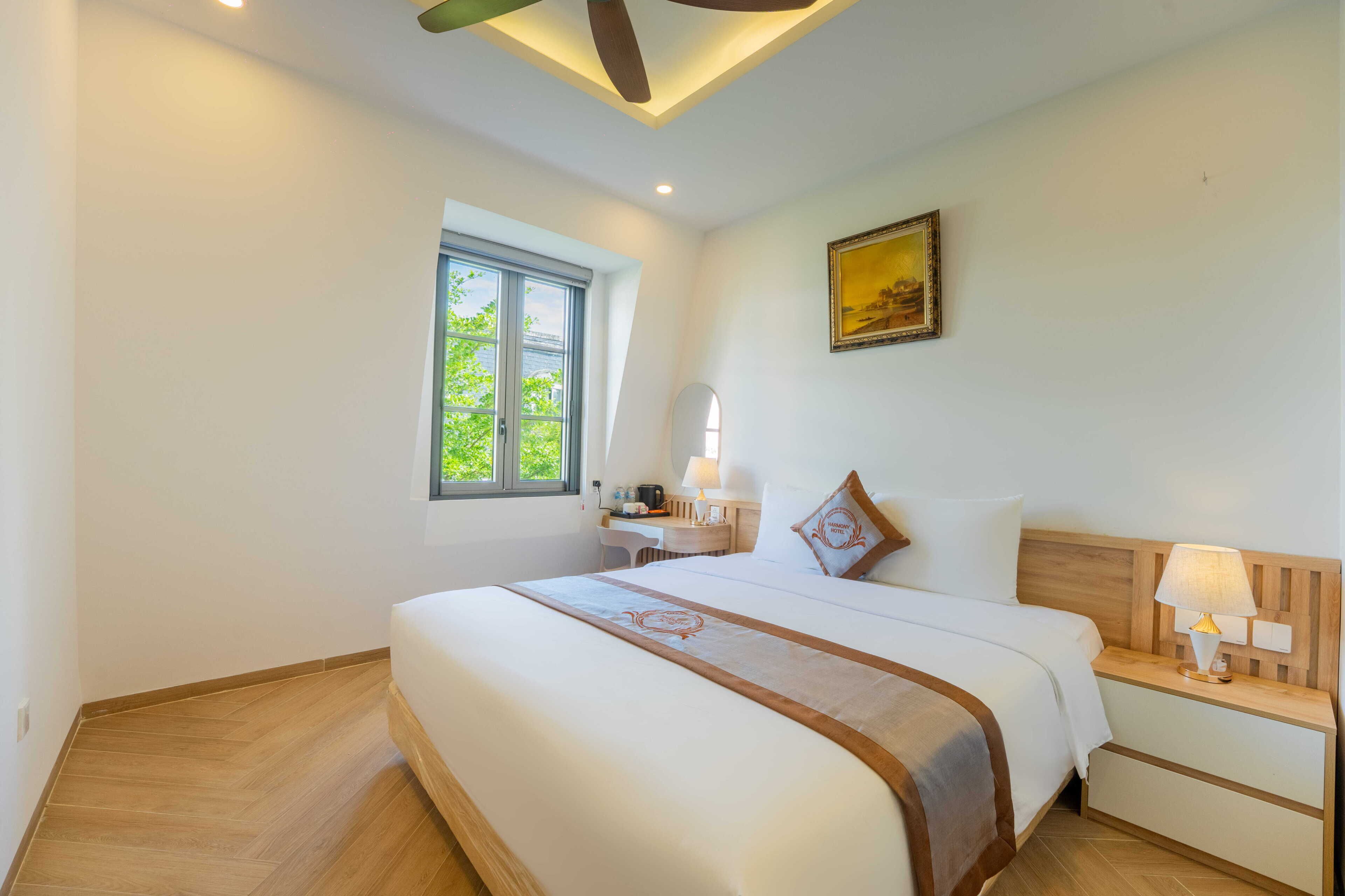 Habitación de hotel con una cama grande, un escritorio, una silla, una mesita de noche con lámpara, una ventana con vistas a zonas verdes y un ventilador de techo.