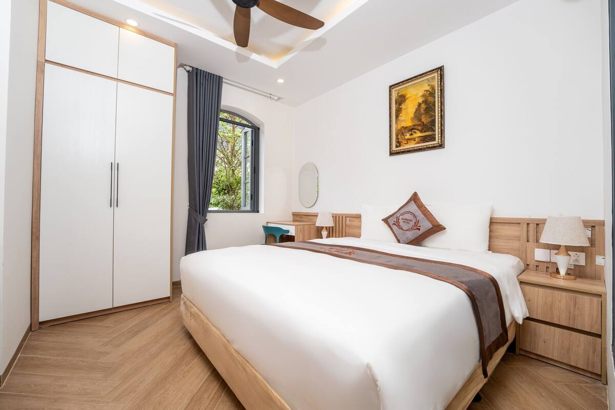 A bedroom with a large bed, a wardrobe, a desk, a chair, a nightstand, a lamp, a ceiling fan, a window with curtains, and a painting on the wall.