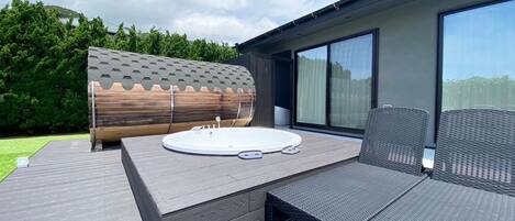 A modern outdoor hot tub area with a wooden deck, a seating area with wicker chairs, and a large window.