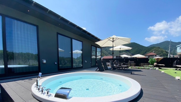 Un bain à remous sur une terrasse en bois, agrémentée de meubles d’extérieur et de parasols, offrant une vue sur un paysage montagneux.