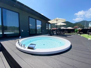 Un bain à remous sur une terrasse en bois, agrémentée de meubles d’extérieur et de parasols, offrant une vue sur un paysage montagneux.