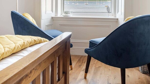 Une pièce avec un lit en bois, deux chaises bleues, une table en bois et une grande fenêtre donnant sur le port.
