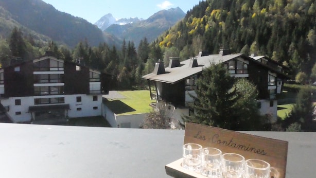 Blick auf Berge mit einem Chalet und einem Schild mit der Aufschrift 'Les Contaminens', sowie einem Tablett mit kleinen Gläsern.