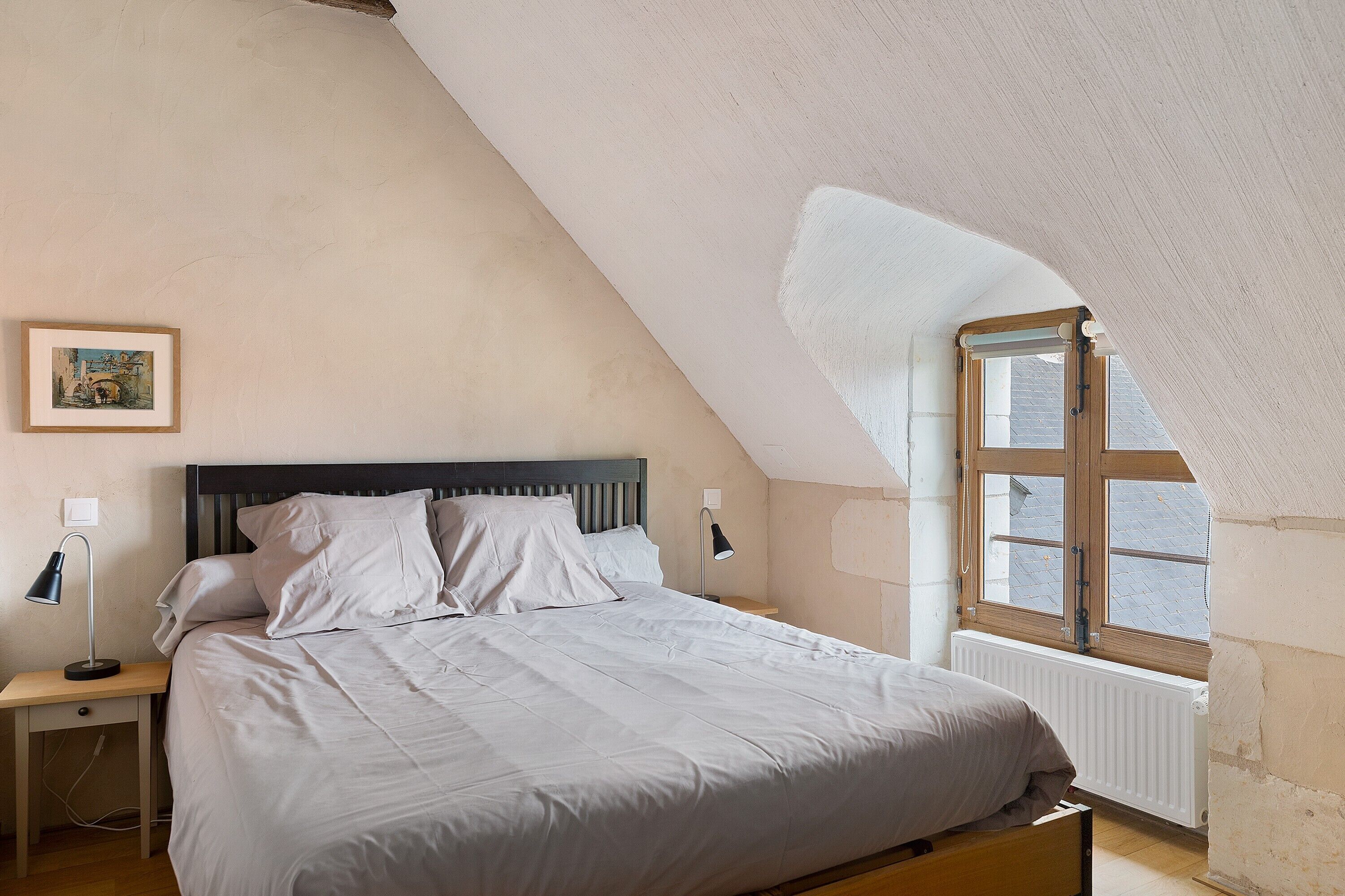 Une chambre avec un plafond en pente, un lit recouvert de linge de lit blanc, une table de chevet avec une lampe et une fenêtre avec des cadres en bois.