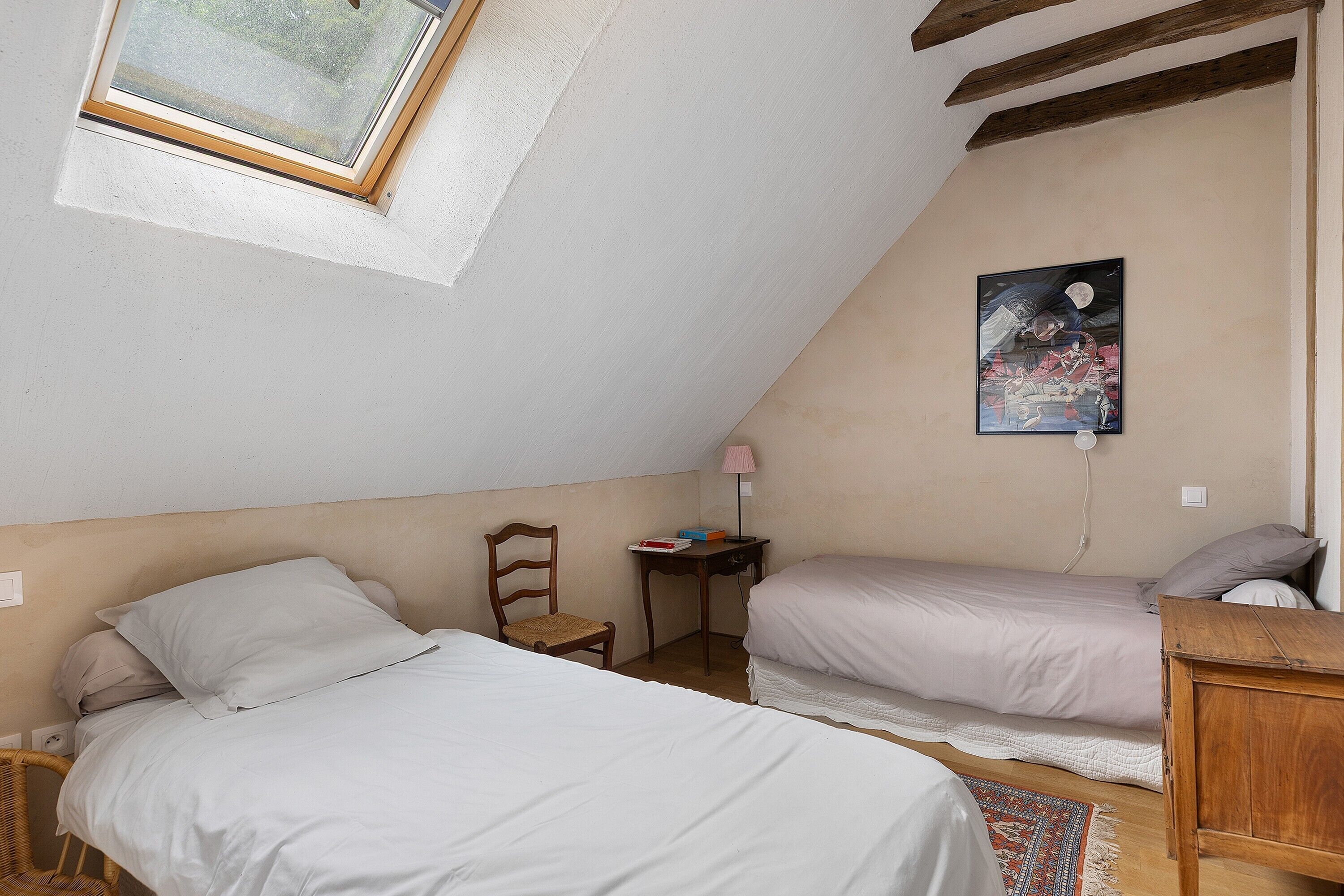 Un dormitorio con dos camas, una silla de madera, una mesita con libros, una lámpara y un cuadro en la pared.