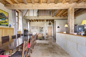 Un comedor rústico con una larga mesa de madera y sillas, una zona de cocina con refrigerador y una lámpara, y un pasillo que conduce a otra habitación.