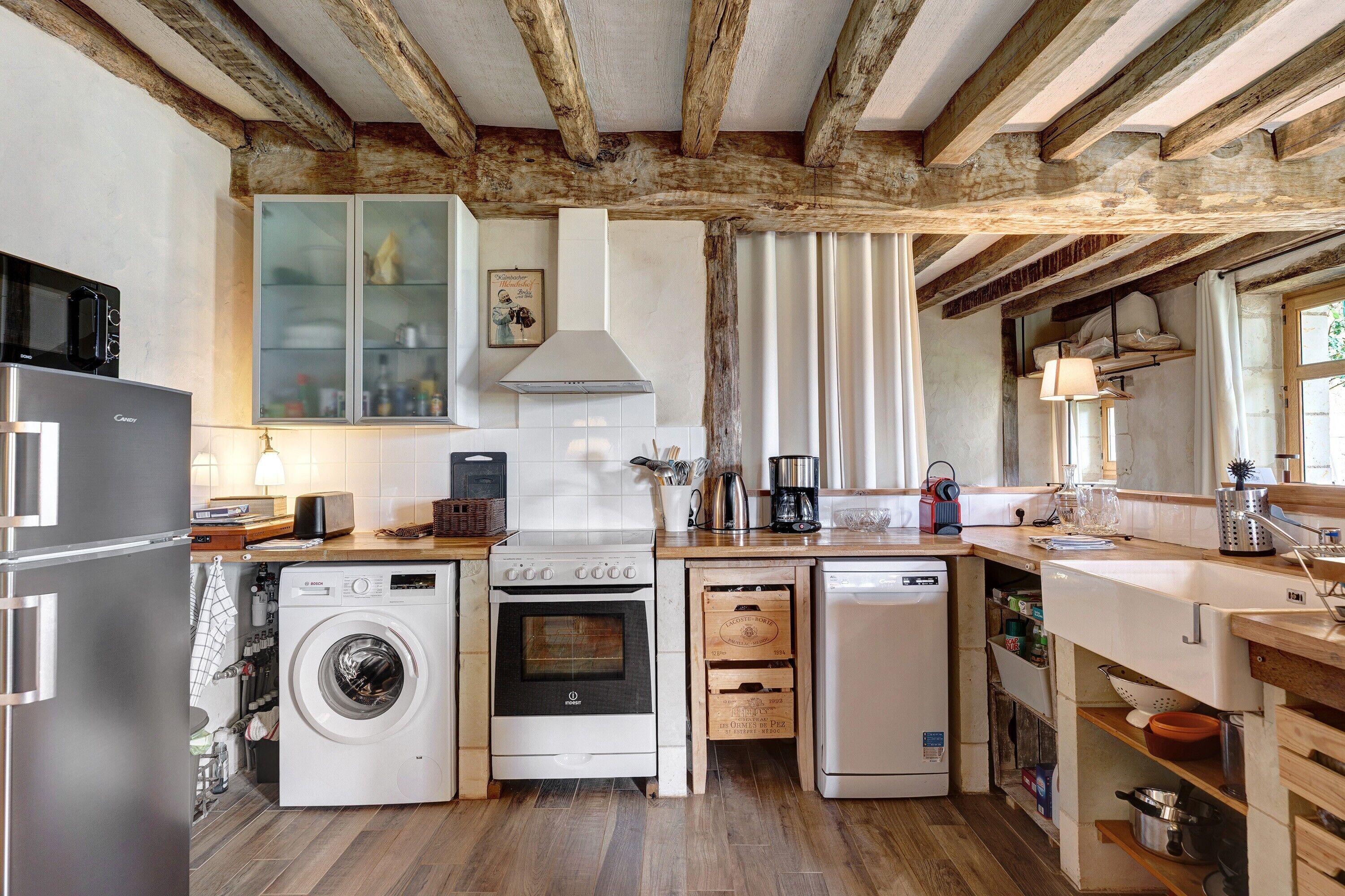Una cucina funzionale con lavatrice, forno e lavastoviglie, travi a vista in legno e una finestra con tende.