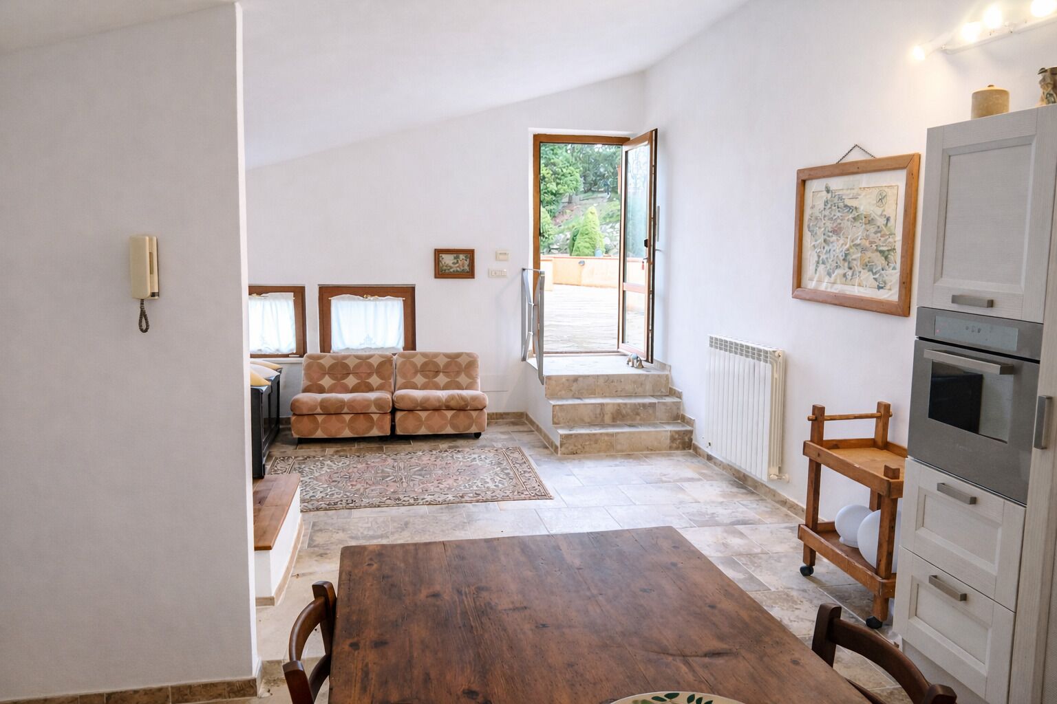 A kitchen with a dining table, a sofa, a staircase leading to a balcony, and a view of the outdoors.