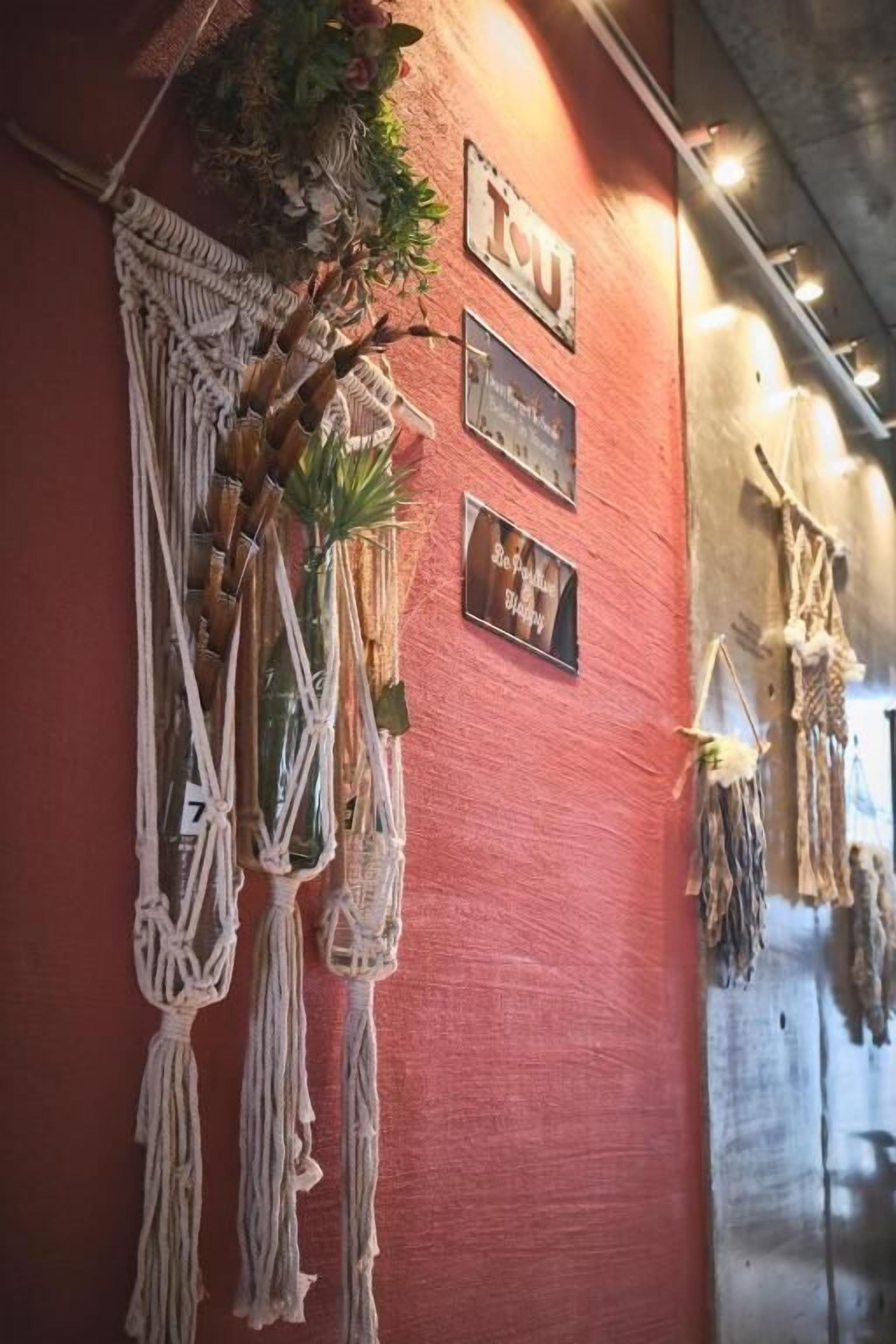 A macrame wall hanging with a plant, a plaque with text, and a metal sign on a red wall.