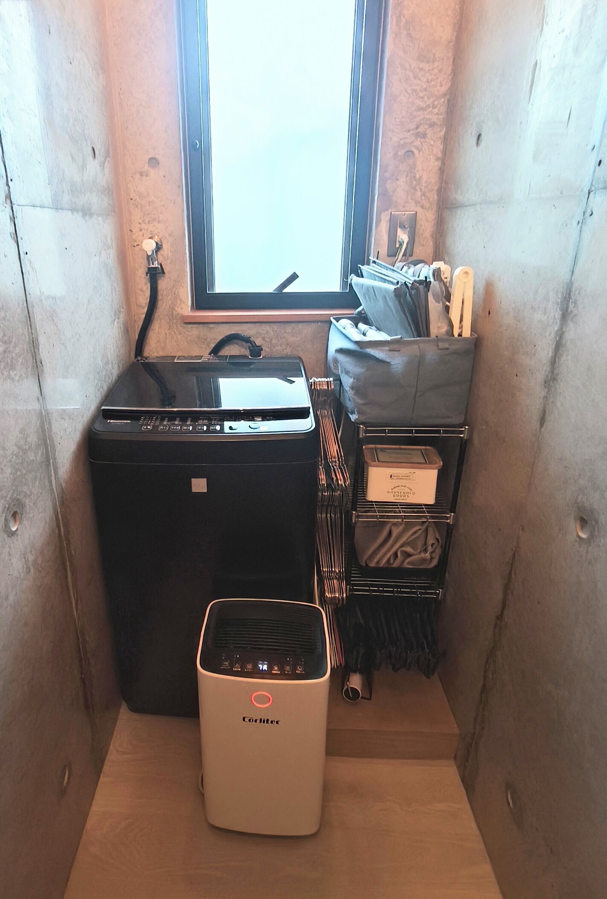 Een kleine kamer met raam, een zwart apparaat, een witte verwarming en een metalen rek met spullen.