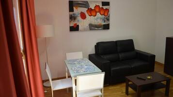 Un salon avec un canapé noir, une petite table, une chaise blanche et un tableau accroché au mur.