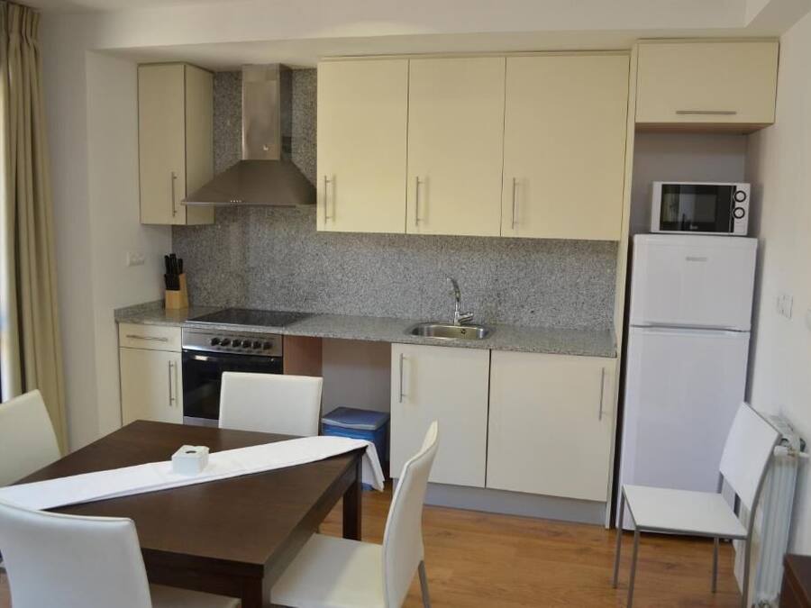 Una cocina compacta con estufa, microondas y refrigerador, una mesa de comedor con sillas, y una ventana con cortinas.