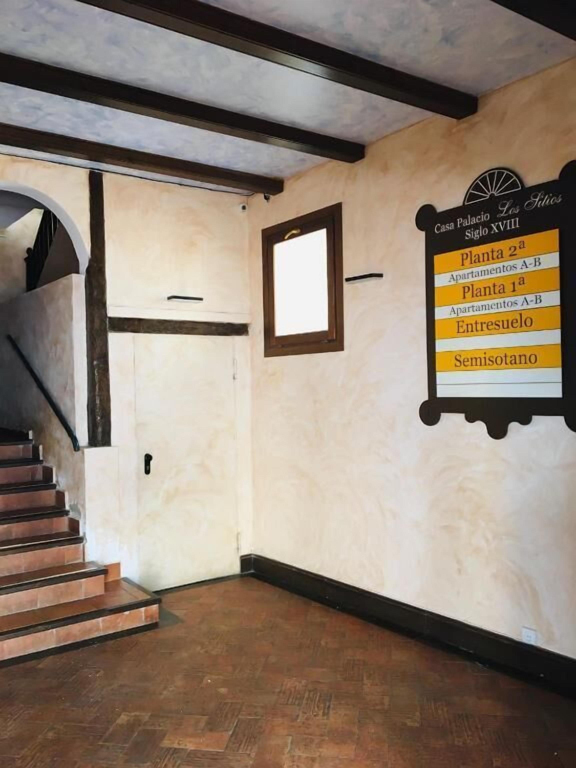Una escalera que lleva a una puerta, un letrero en la pared con nombres de habitaciones, y una ventana con vistas.