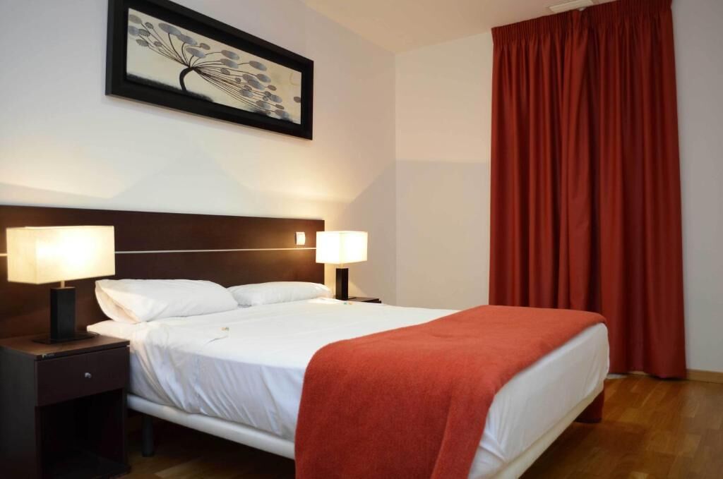 Ett hotellrum med en säng, två sänglampor, en inramad tavla på väggen och en röd gardin.