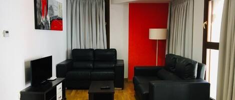 Una sala de estar con un sofá negro, una mesita, un televisor sobre un mueble bajo y una pared de acento roja.