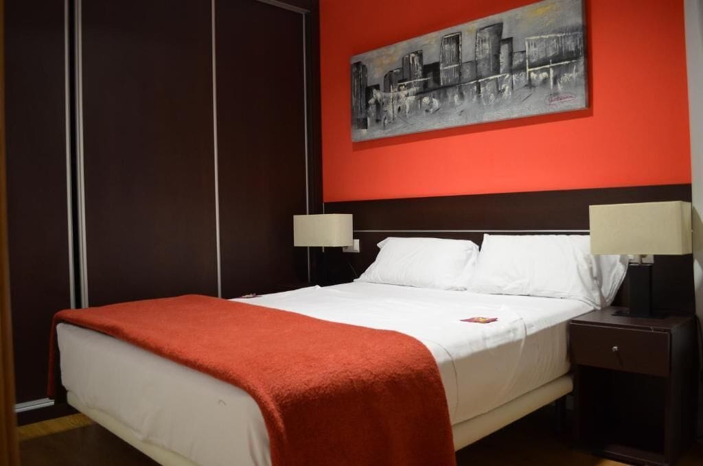 A hotel room with a bed, two bedside tables with lamps, a red accent wall, and a cityscape painting.