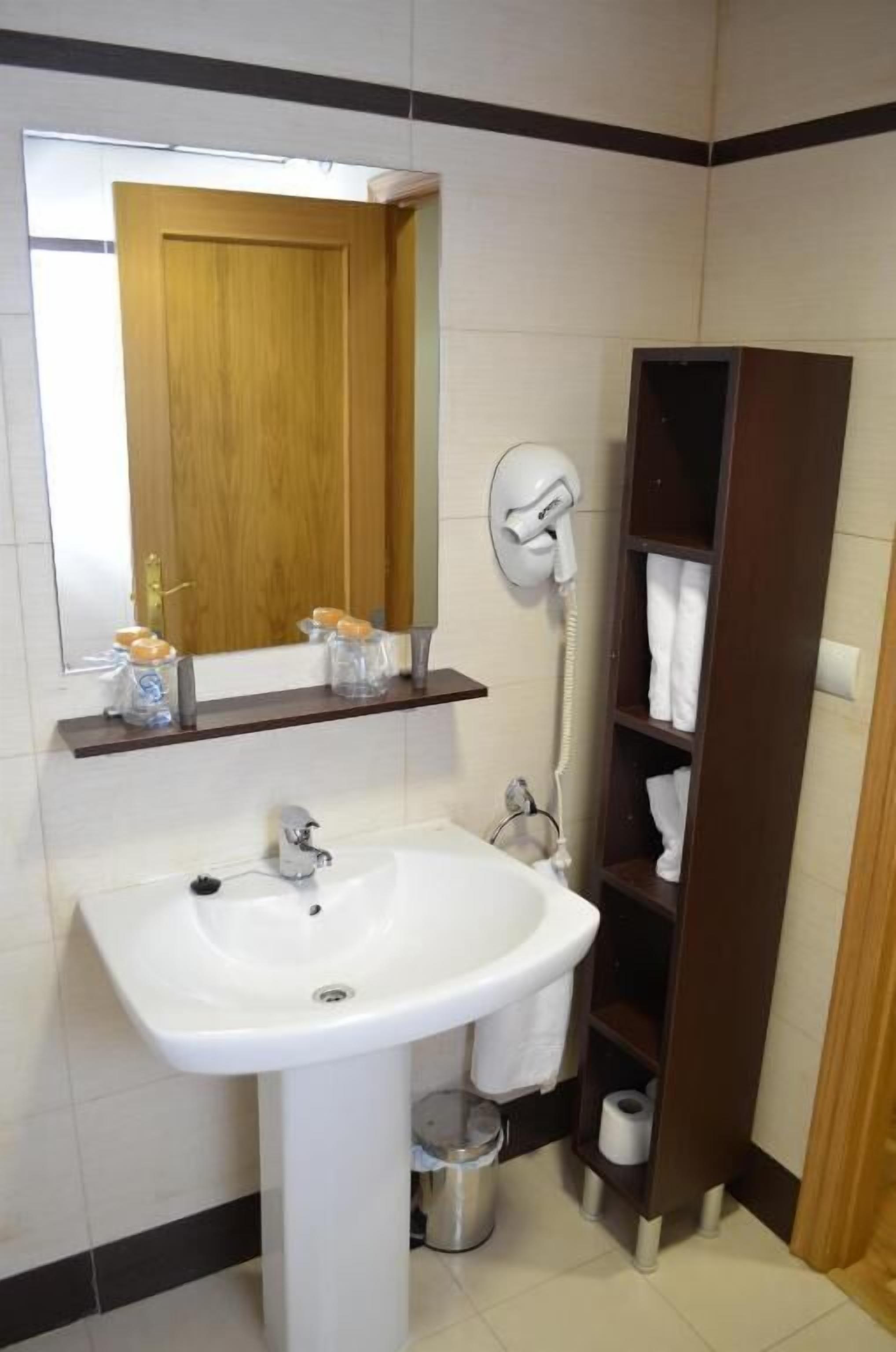 Un baño con lavamanos blanco, un espejo, una puerta de madera y un estante con artículos de aseo.