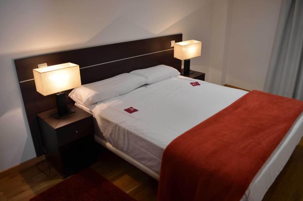 Habitación de hotel con cama, dos lámparas de noche, mesita de noche y una manta roja.