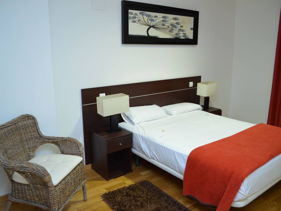 A hotel room with a bed, two bedside lamps, a wicker chair, a nightstand, a red blanket, a brown rug, and a framed picture on the wall.