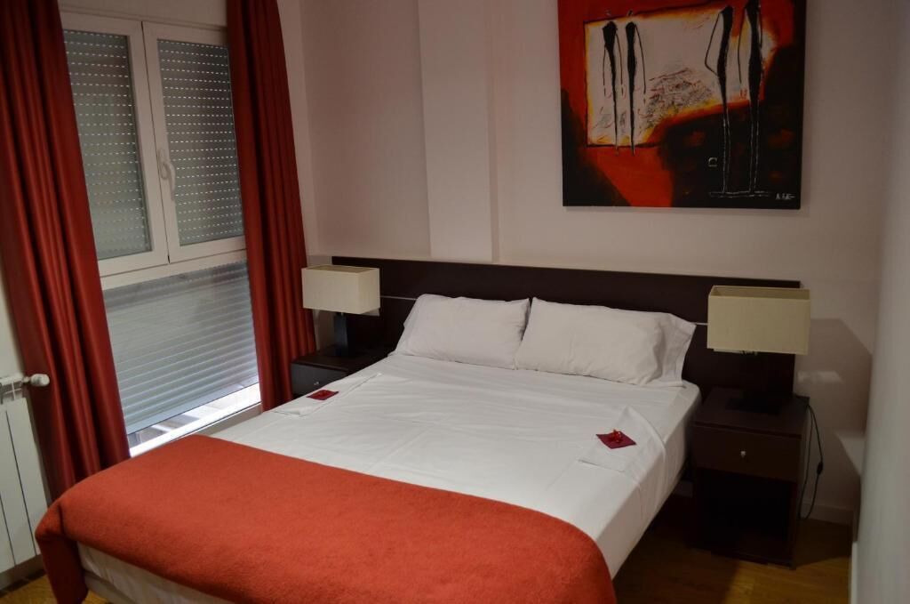 Habitación de hotel con cama, dos lámparas de noche, una ventana con cortinas y un cuadro en la pared.
