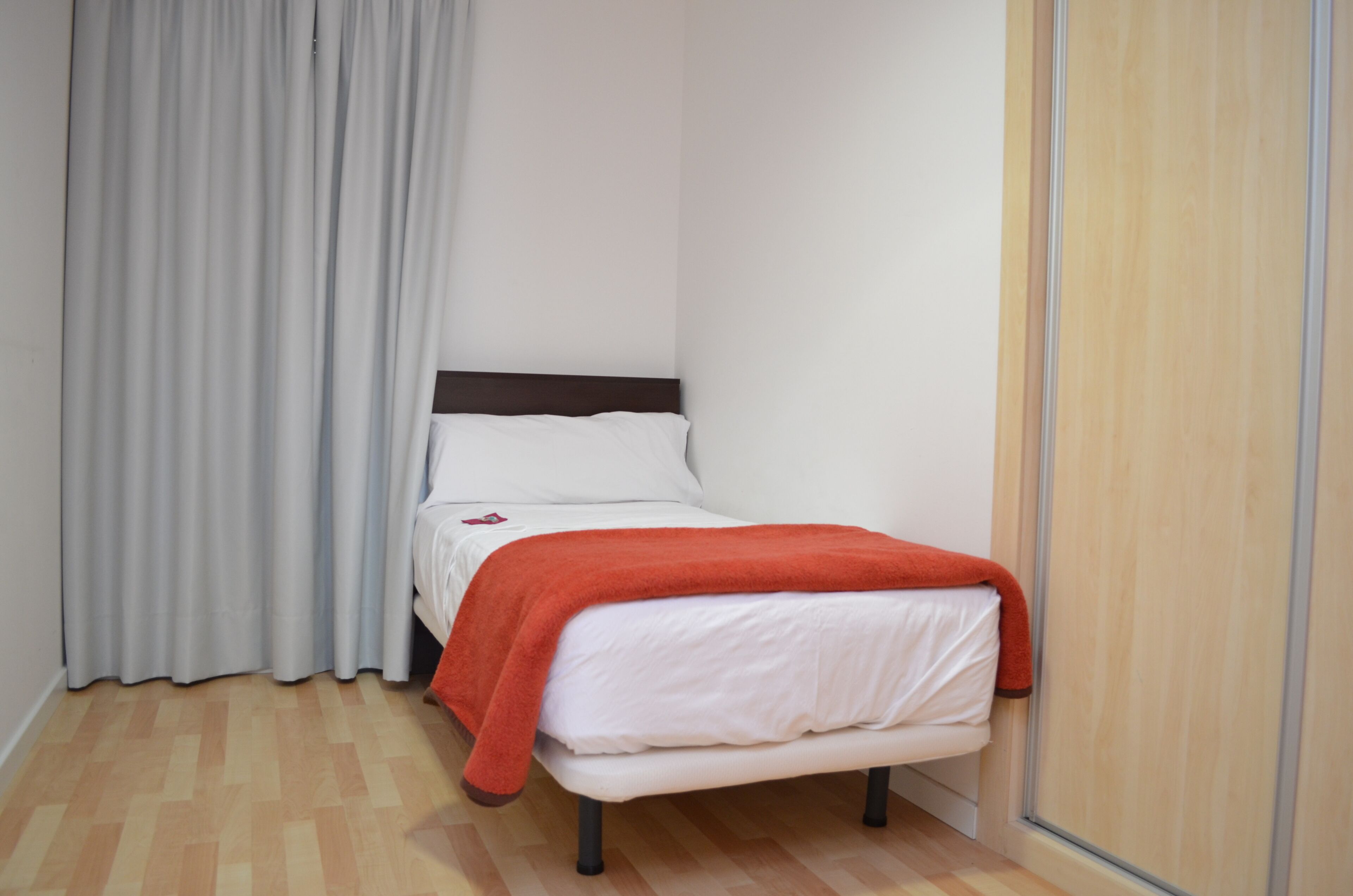 A small, neatly arranged bedroom with a single bed, a wooden floor, and a grey curtain.
