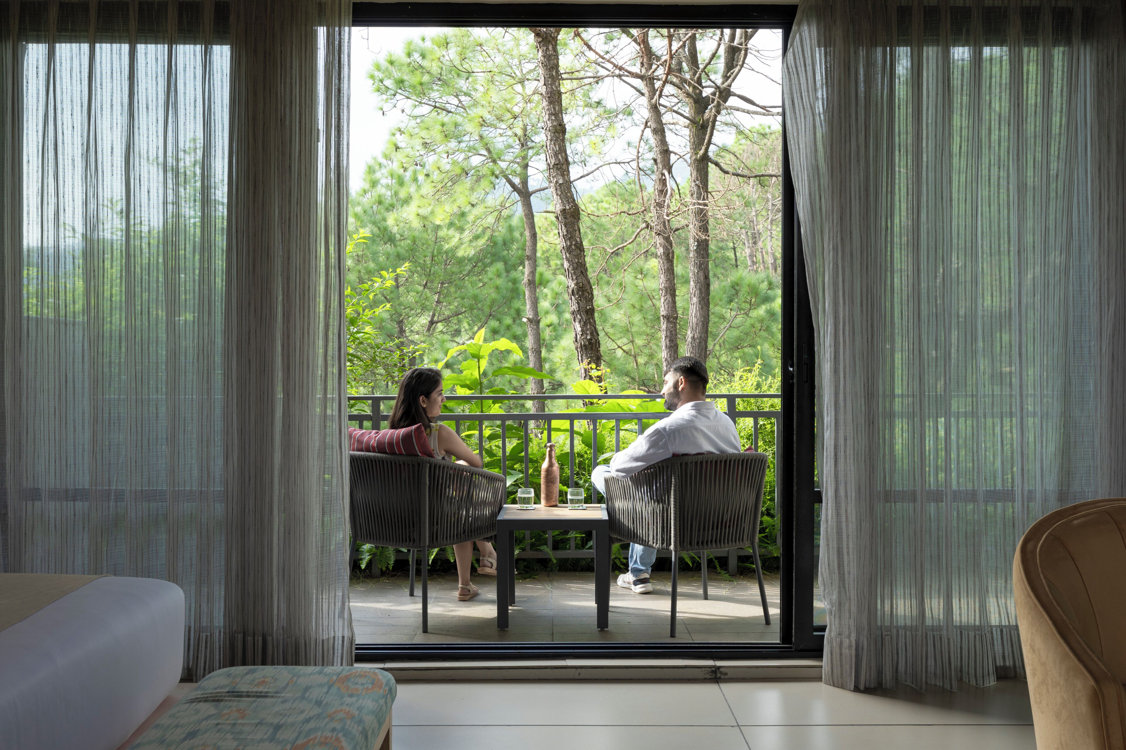 Una habitación con una cama, una mesa con dos sillas y vistas a un balcón con barandilla y vegetación.