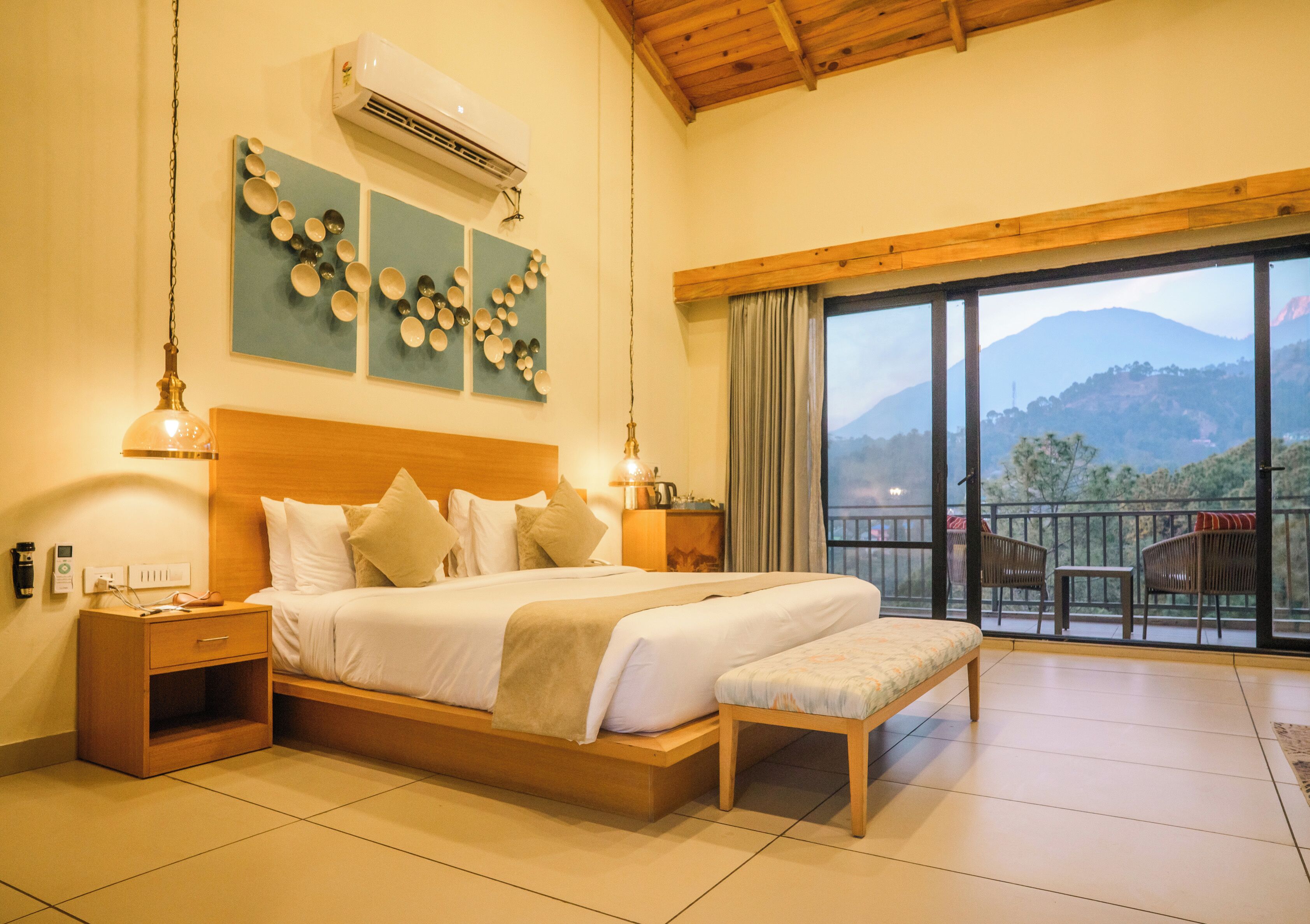 Habitación de hotel con una cama grande, mesitas de noche, un banco, vistas a las montañas y un balcón con sillas.