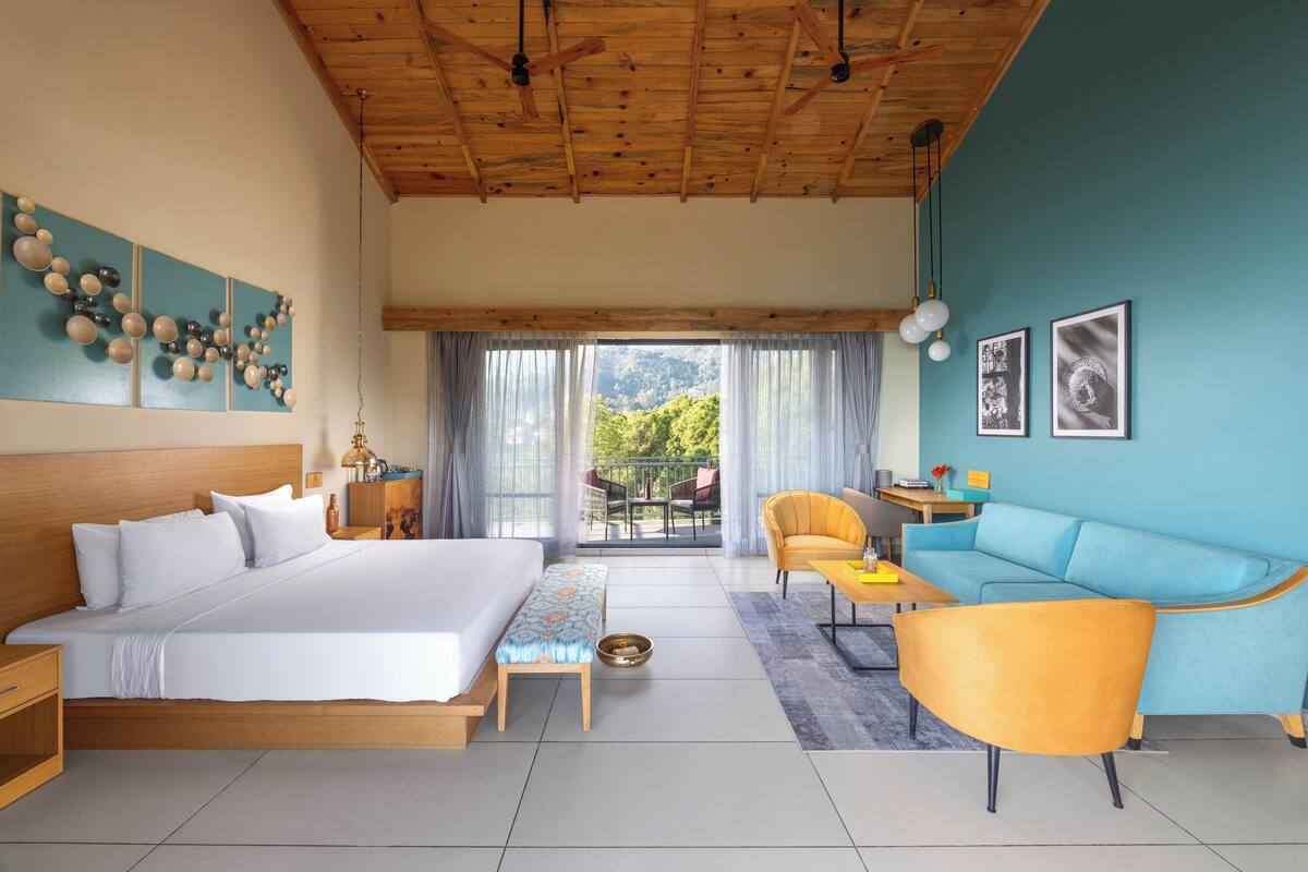 A room with a bed, a sofa, a dining area, and a view of greenery.