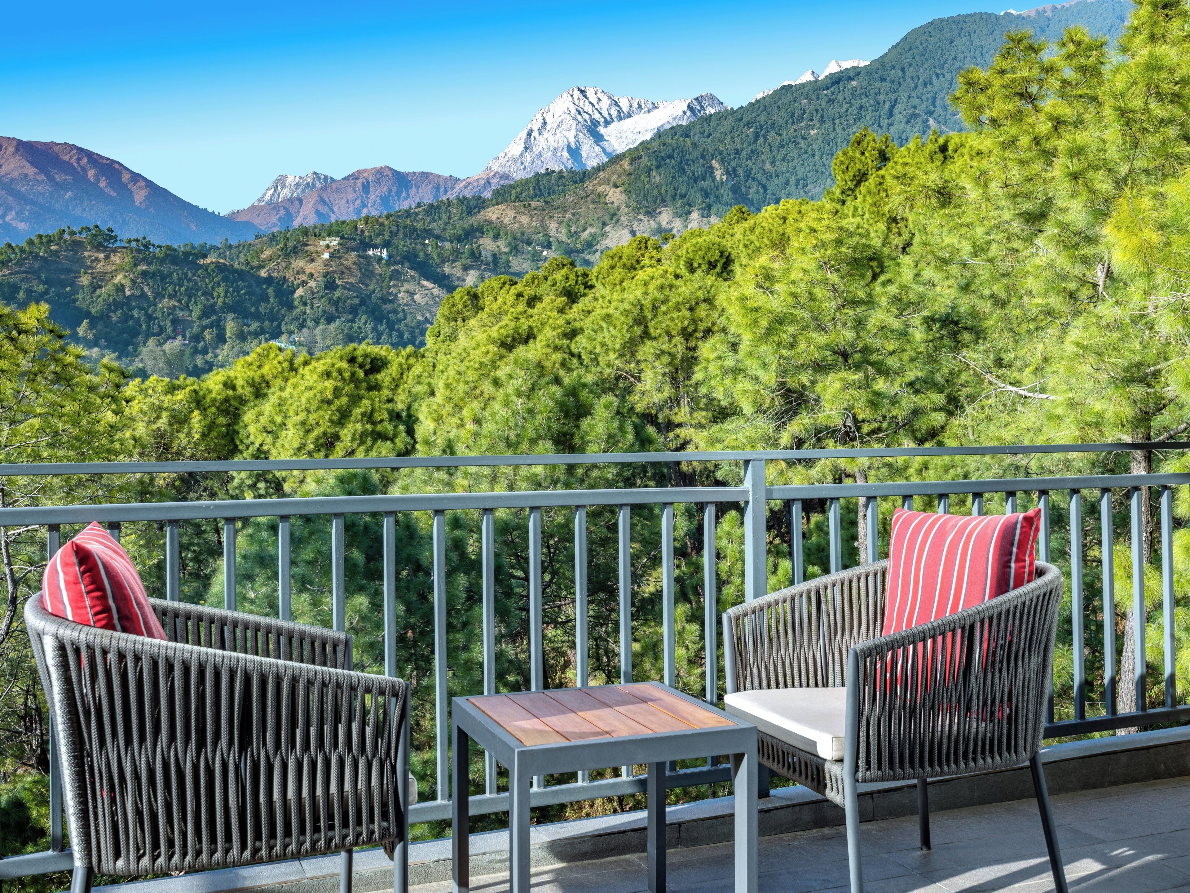 A balcony with wicker chairs and a small table, overlooking a mountainous landscape with pine trees.