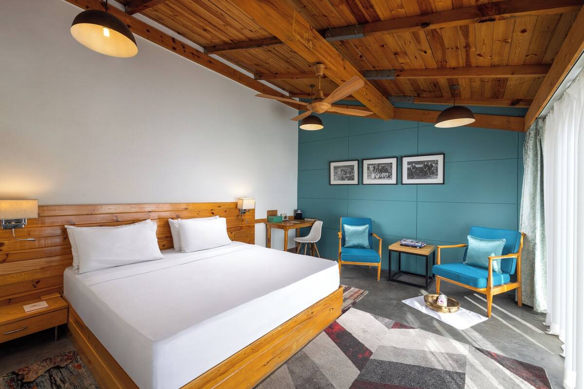 A hotel room with a wooden ceiling, a large bed, a desk, two chairs, and a wall with framed pictures.