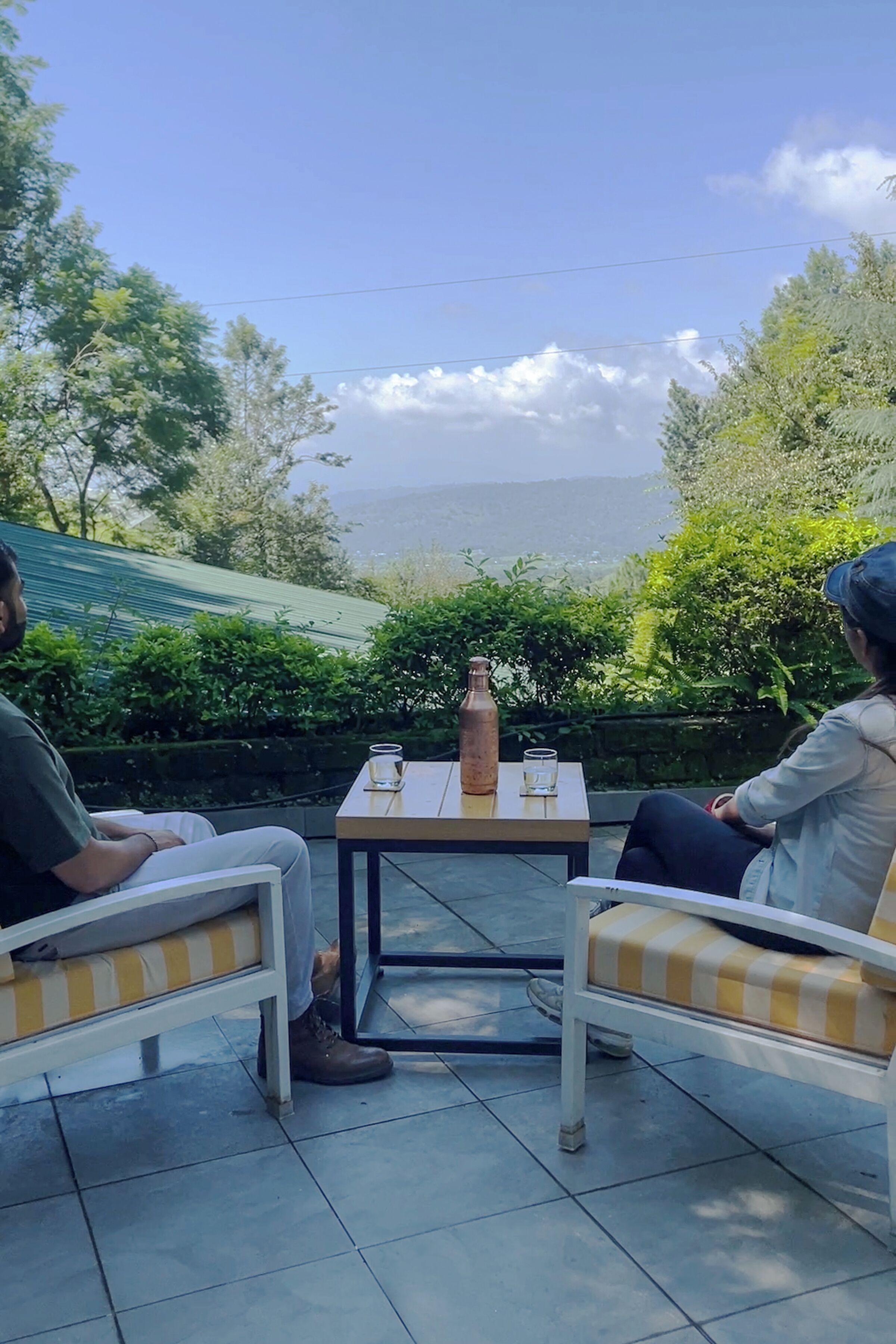 Two people seated at a table with a bottle and glasses, outdoors with a view of trees and hills.