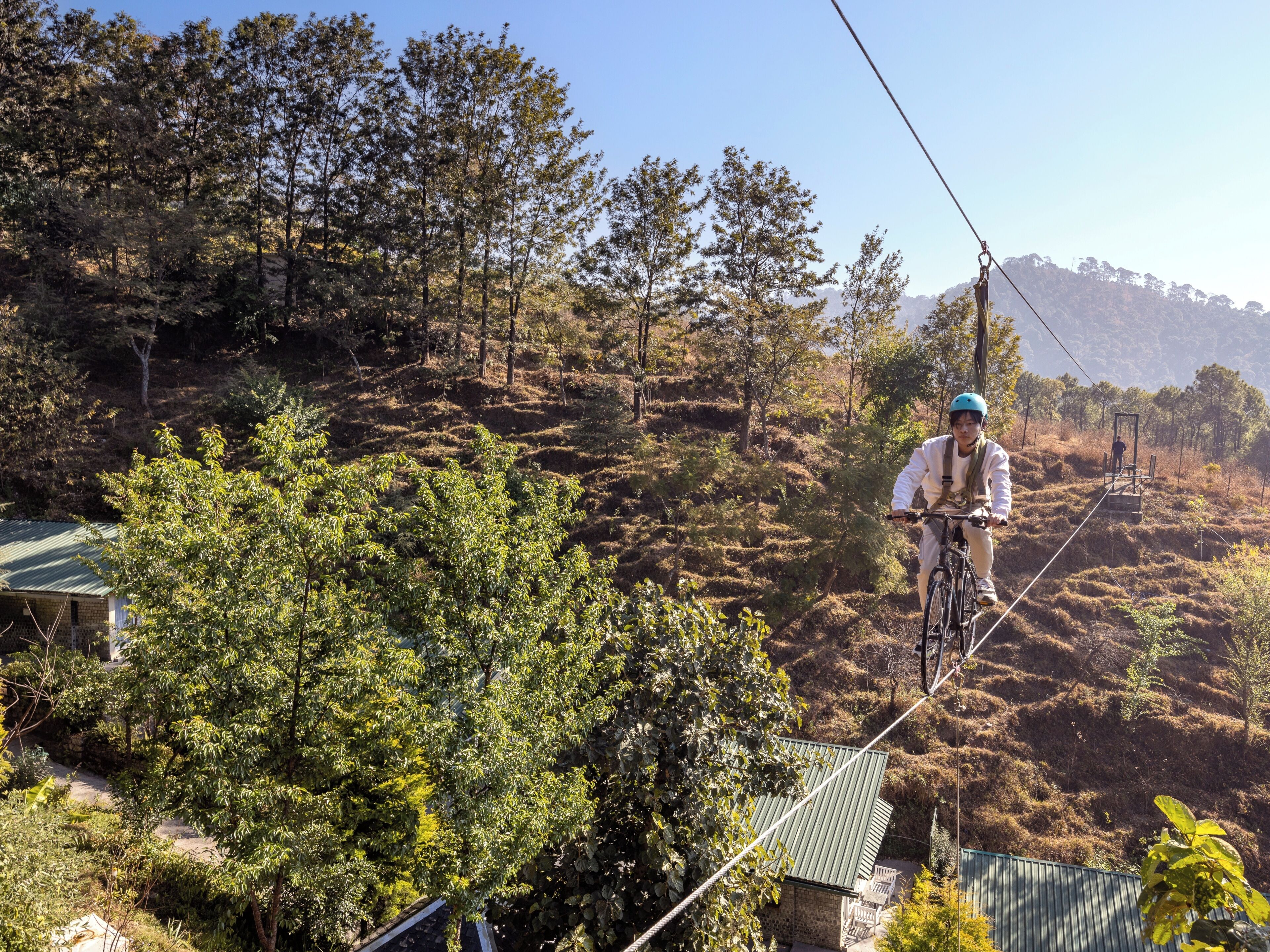 A person on a bicycle on a rope bridge in a hilly area with trees and a building with a green roof.