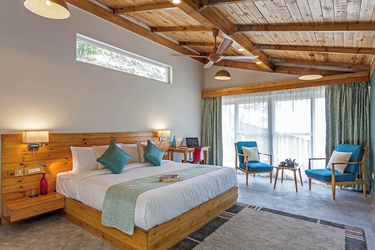 A bedroom with a wooden ceiling, a bed with pillows, a desk with a laptop, and two chairs.
