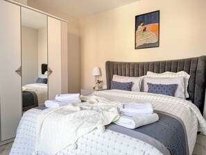 Ein ordentlich bezogenes Bett mit grauem Kopfteil, einer Nachttischlampe und einem Bild an der Wand.