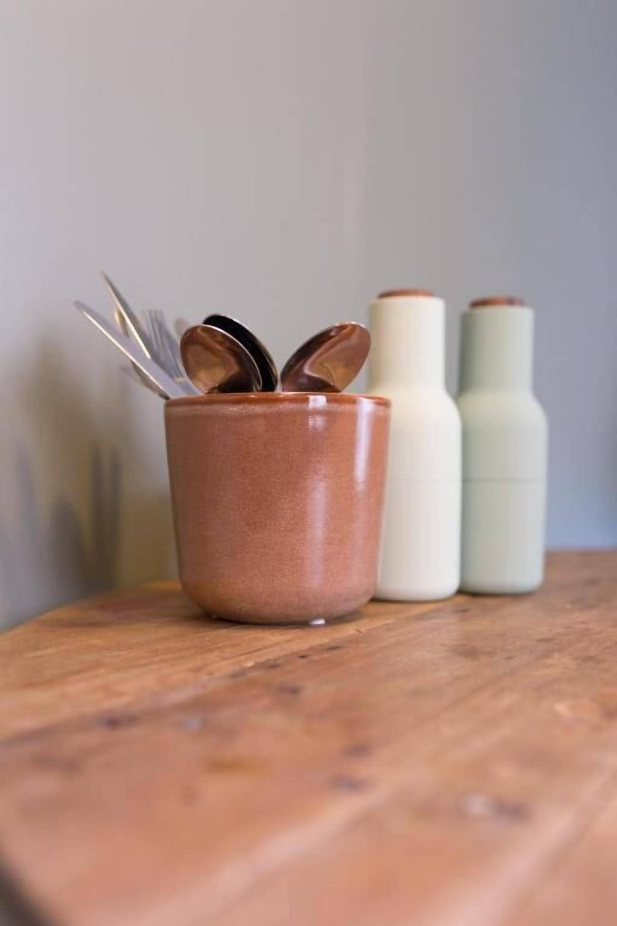 A wooden surface with a copper-colored mug containing spoons and two white bottles with brown caps.