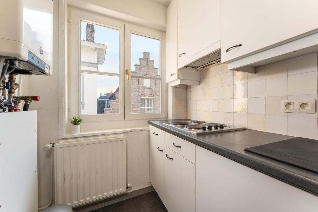 Een keuken met witte kastjes, een fornuis, een raam met uitzicht op gebouwen, en een radiator.